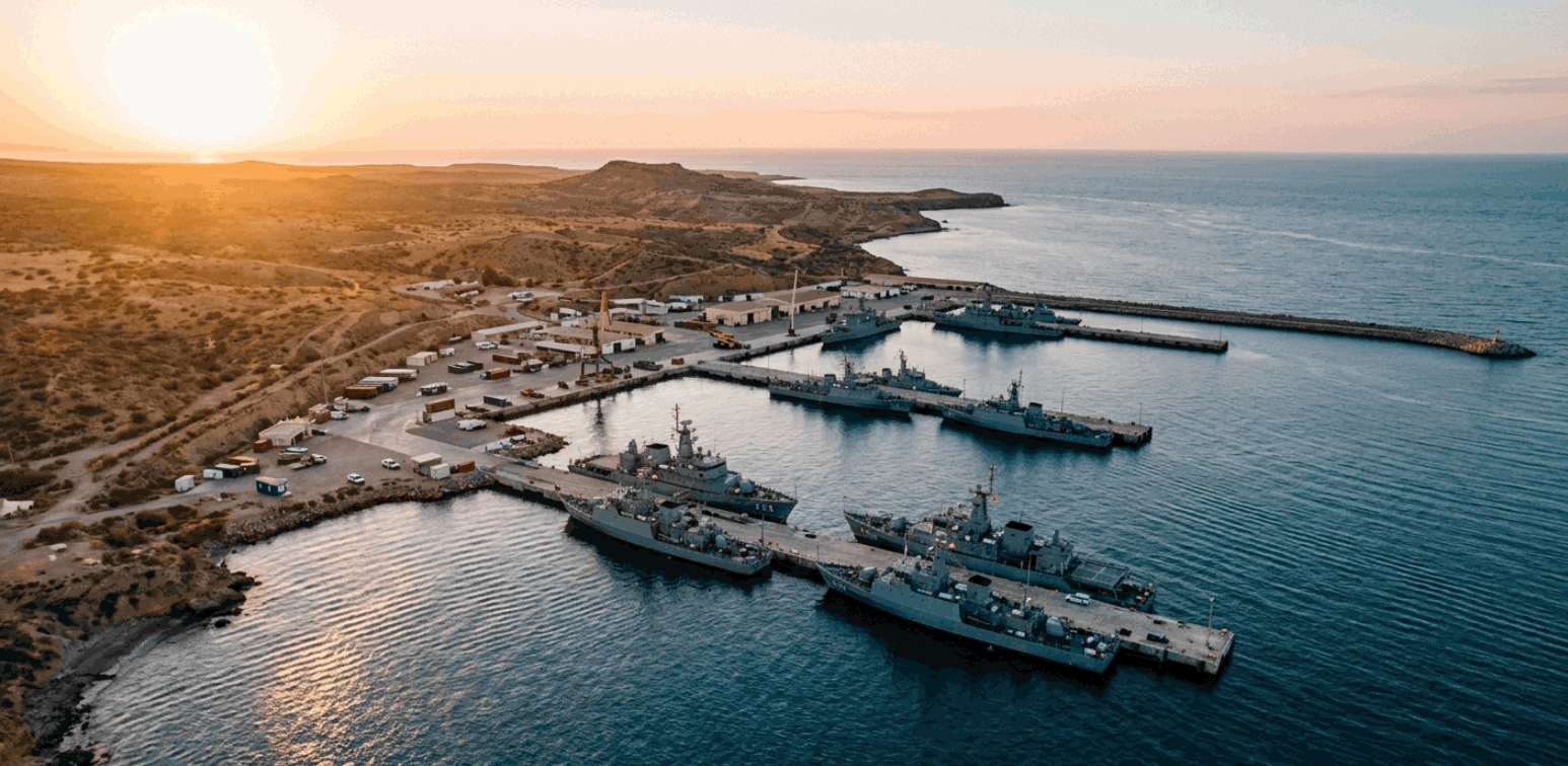 Naval base on an arid Gulf coastline at golden hour — grey warships at dock, the military architecture of chokepoint sovereignty