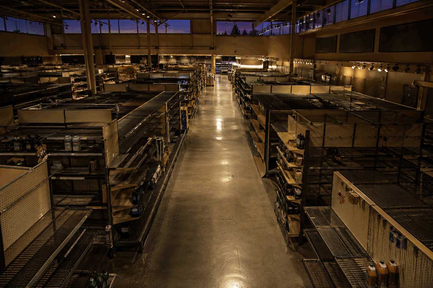 Supermarket aisle interior at end of day, half-empty shelves and bare bays in sodium light, dusk through high clerestory windows — the household squeeze made literal