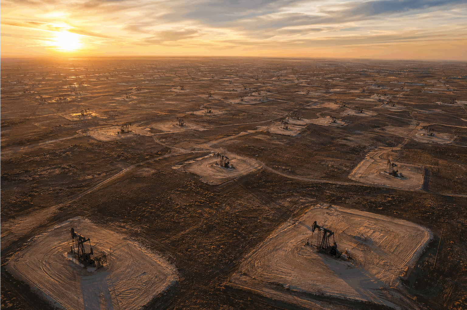 Drone aerial of a vast shale oil basin at sunset, dozens of pump jacks scattered across cleared well pads in flat semi-arid terrain stretching to horizon — geology binds, no announcement changes the rig count